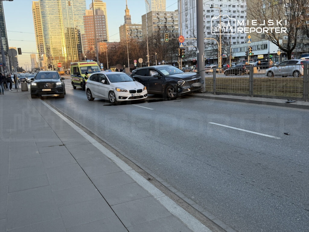 Dwa uszkodzone samochody osobowe na jezdni w miejskim otoczeniu, obok przechodniów i budynków w tle.