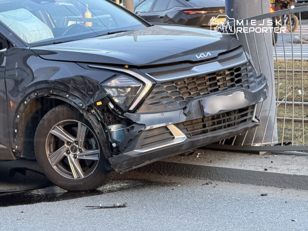 Czarny samochód osobowy z uszkodzoną przednią częścią stoi na poboczu, oparty o ogrodzenie.
