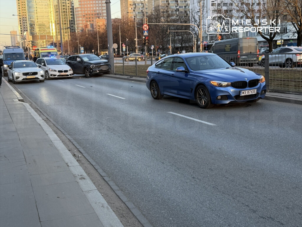 Na jezdni stoją trzy samochody osobowe, w tym niebieskie BMW i czarny pojazd z uszkodzeniami, obok przejeżdżają inne auta.