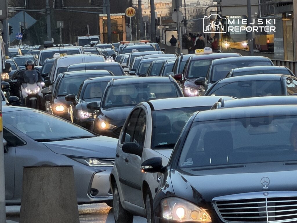 Gęsty ruch samochodowy na ulicy, z licznymi pojazdami w korku, w tle widoczne budynki i znaki drogowe.