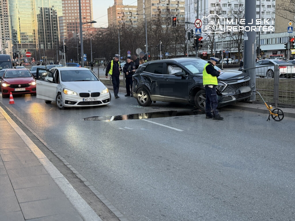 Na ulicy zatrzymane są dwa uszkodzone samochody, a policjanci badają miejsce wypadku obok studzienki.