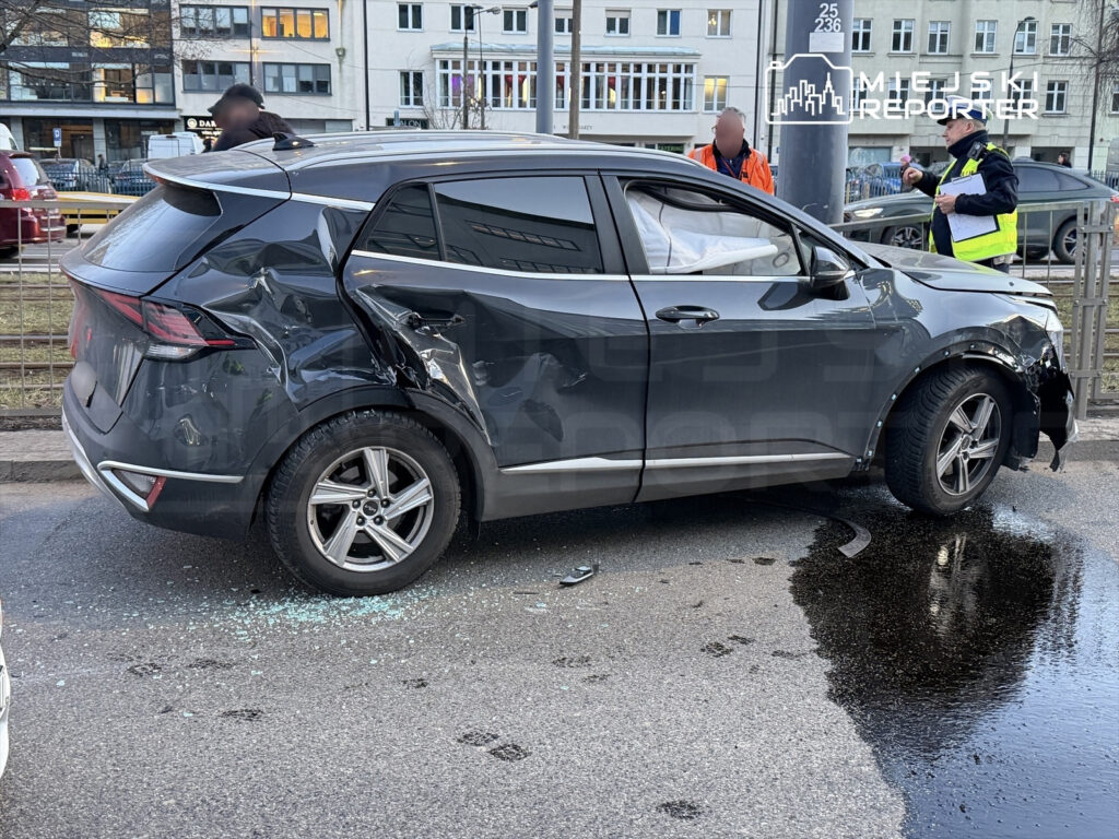Czarny SUV z uszkodzoną prawą stroną stoi na ulicy, obok grupy osób w odzieży roboczej oraz szkła rozbitego na jezdni.
