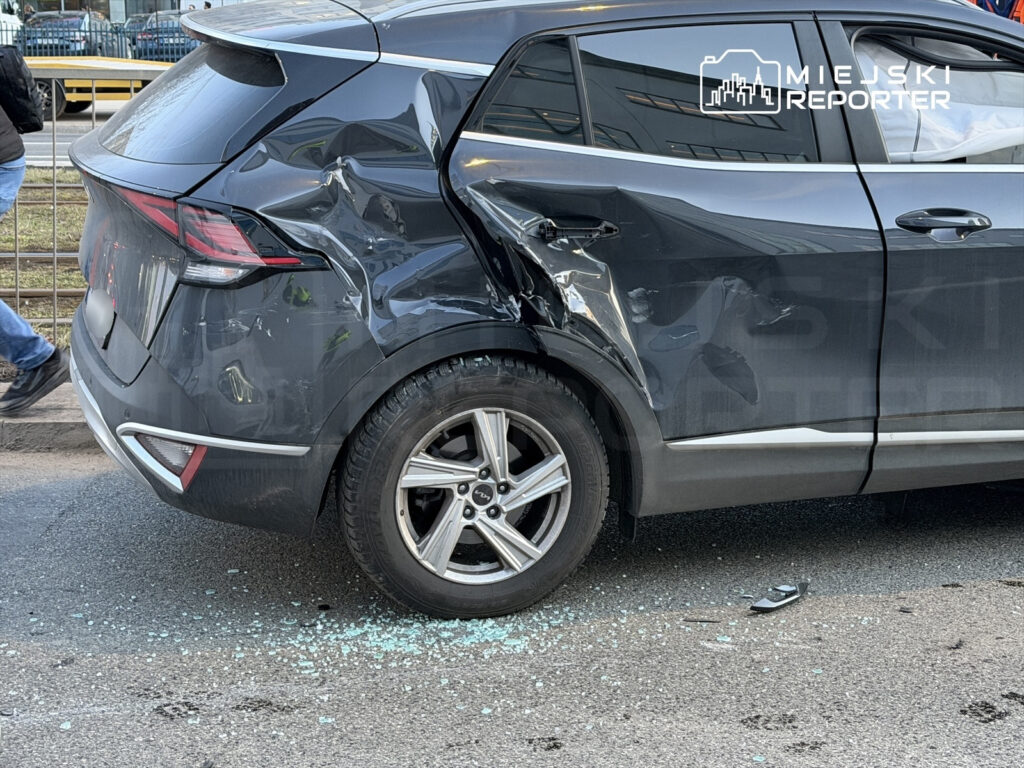 Czarny samochód osobowy z uszkodzeniem tyłu, obok leżą szkła na drodze, w tle przechodzący pieszy.