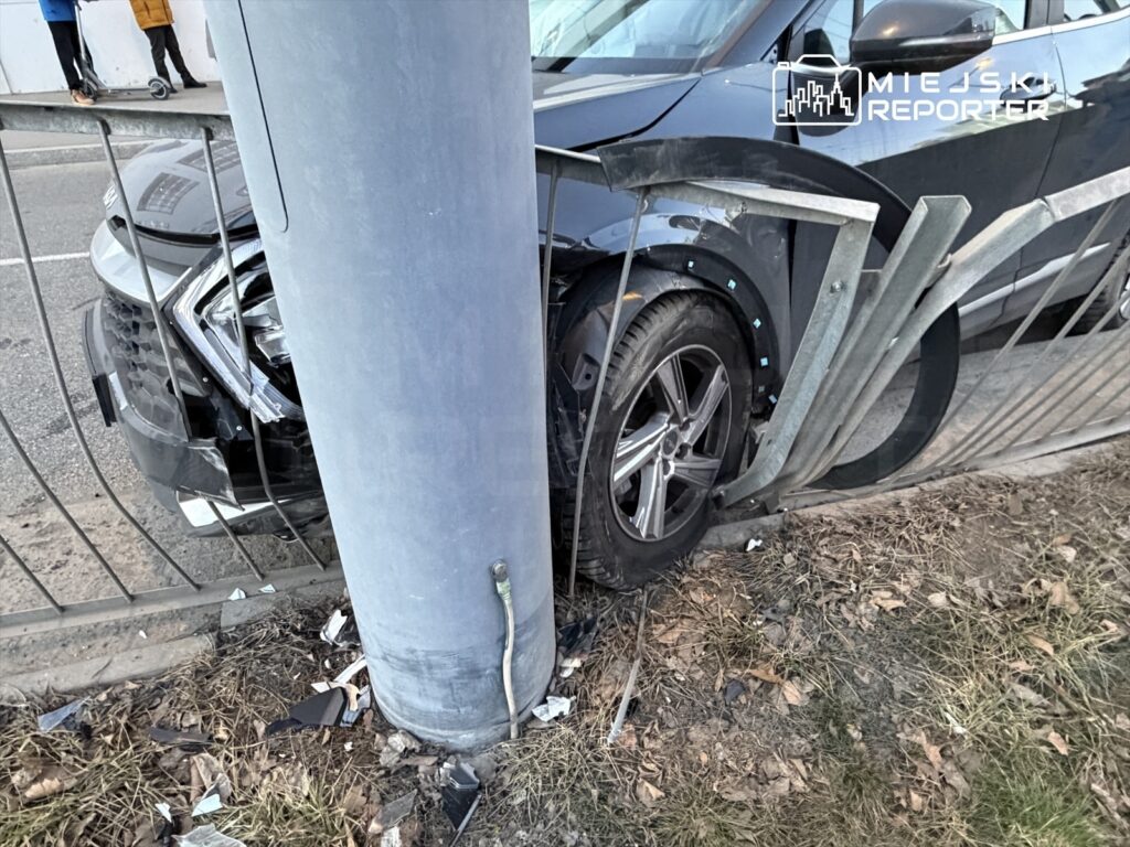 Czarny samochód osobowy wjeżdżający w słup oraz ogrodzenie, z widocznymi uszkodzeniami przodu pojazdu.
