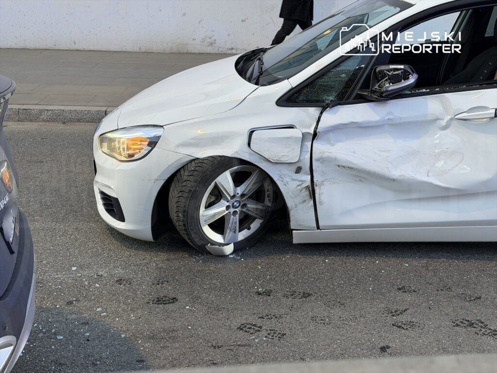 Biały samochód osobowy z uszkodzoną boczną częścią i zerwaną oponą stoi na jezdni. W tle widoczny fragment innego pojazdu.
