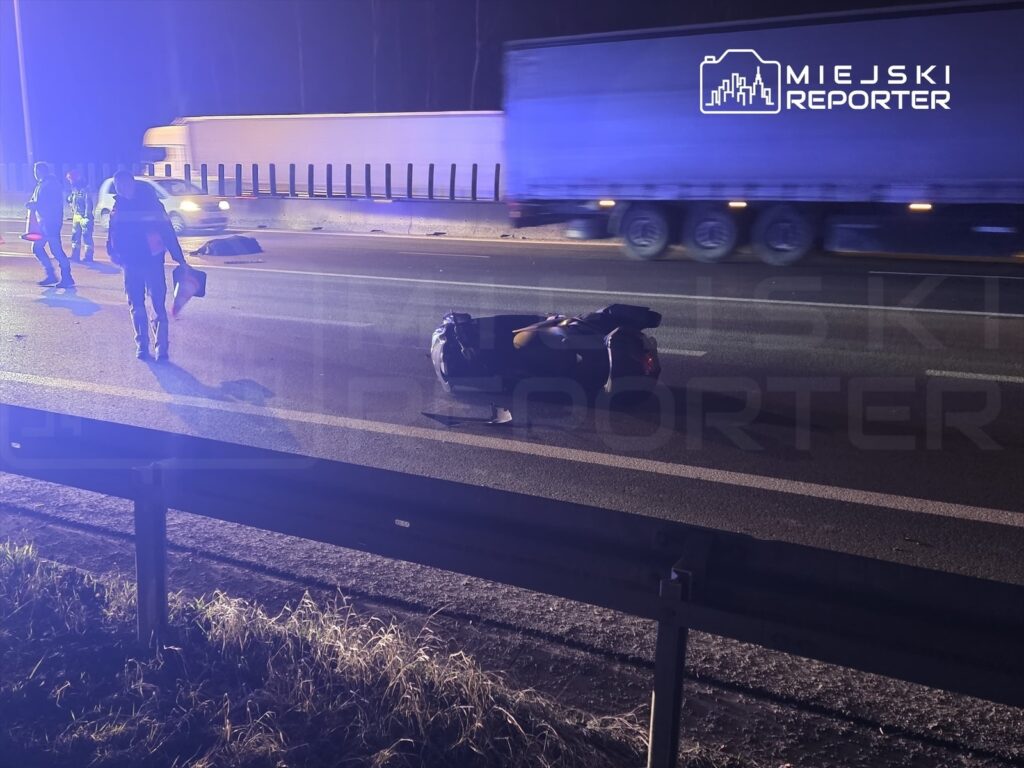 Grupa ratowników na drodze ekspresowej, obok przewróconego motocykla, w tle przejeżdżające ciężarówki.