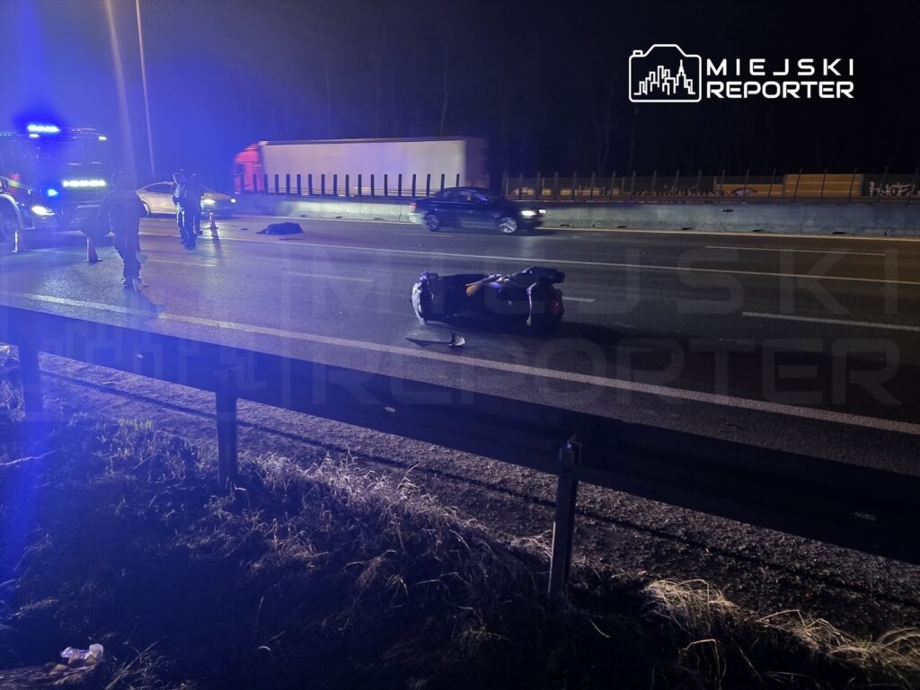 Na drodze ekspresowej leży przewrócony motocykl, a obok pracują ratownicy i policjanci w odblaskowych kamizelkach.