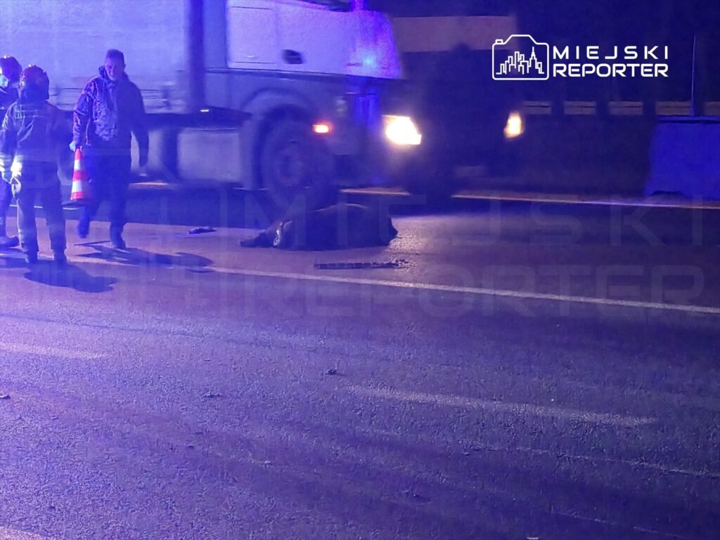 Grupa ratowników w kamizelkach odblaskowych udziela pomocy na jezdni, obok leżącego mężczyzny w nocy.