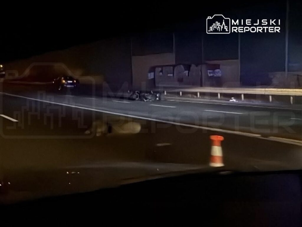 Na drodze widoczny jest wypadek – uszkodzony motocykl leży na jezdni, a obok stoi pachołek drogowy.