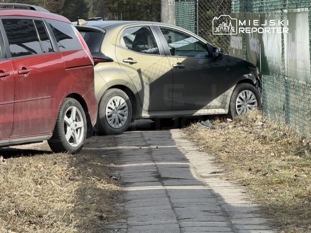 Zielony samochód zaparkowany nieprawidłowo na chodniku obok czerwonego pojazdu, w otoczeniu ogrodzenia.
