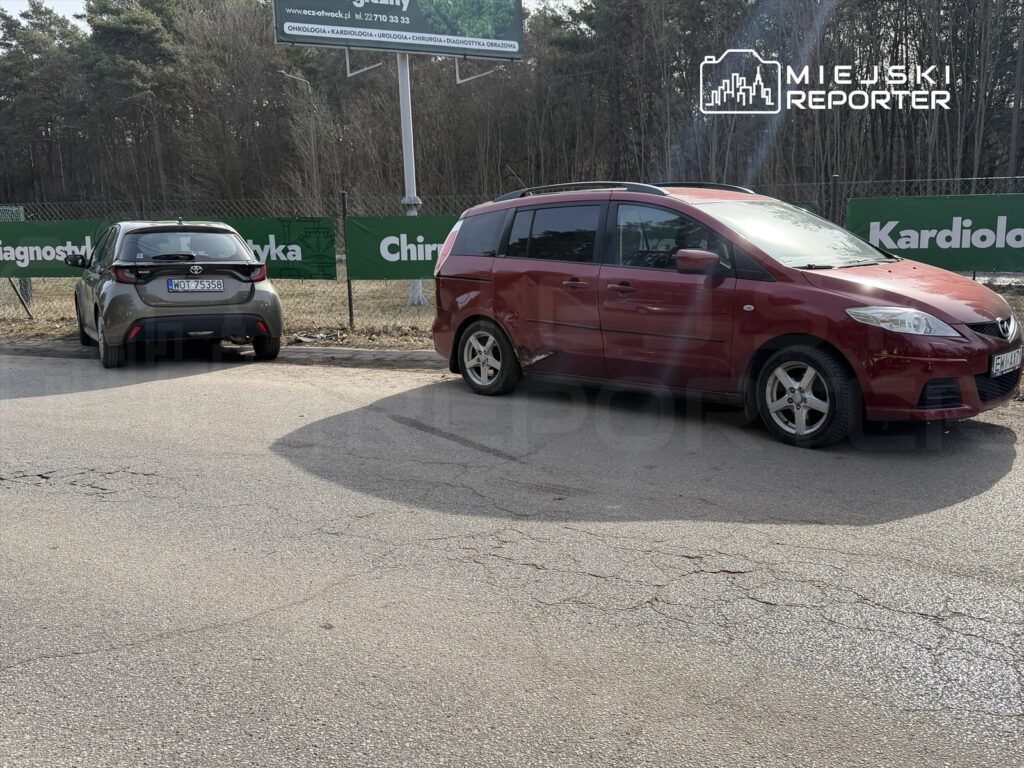 Dwa samochody zaparkowane na poboczu drogi, w tle billboard z informacjami o usługach medycznych.