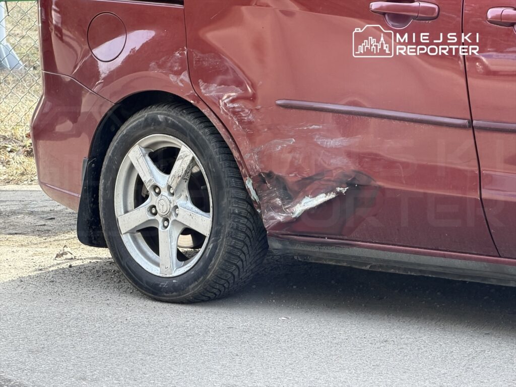 Czerwony samochód z uszkodzoną boczną częścią oraz zarysowaną felgą stoi na drodze.