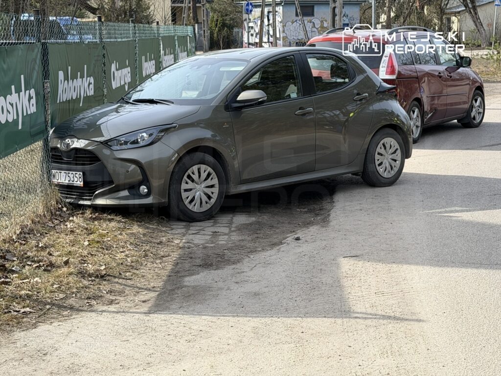 Szary samochód osobowy zaparkowany blisko ogrodzenia w terenie miejskim, obok czerwonego pojazdu.