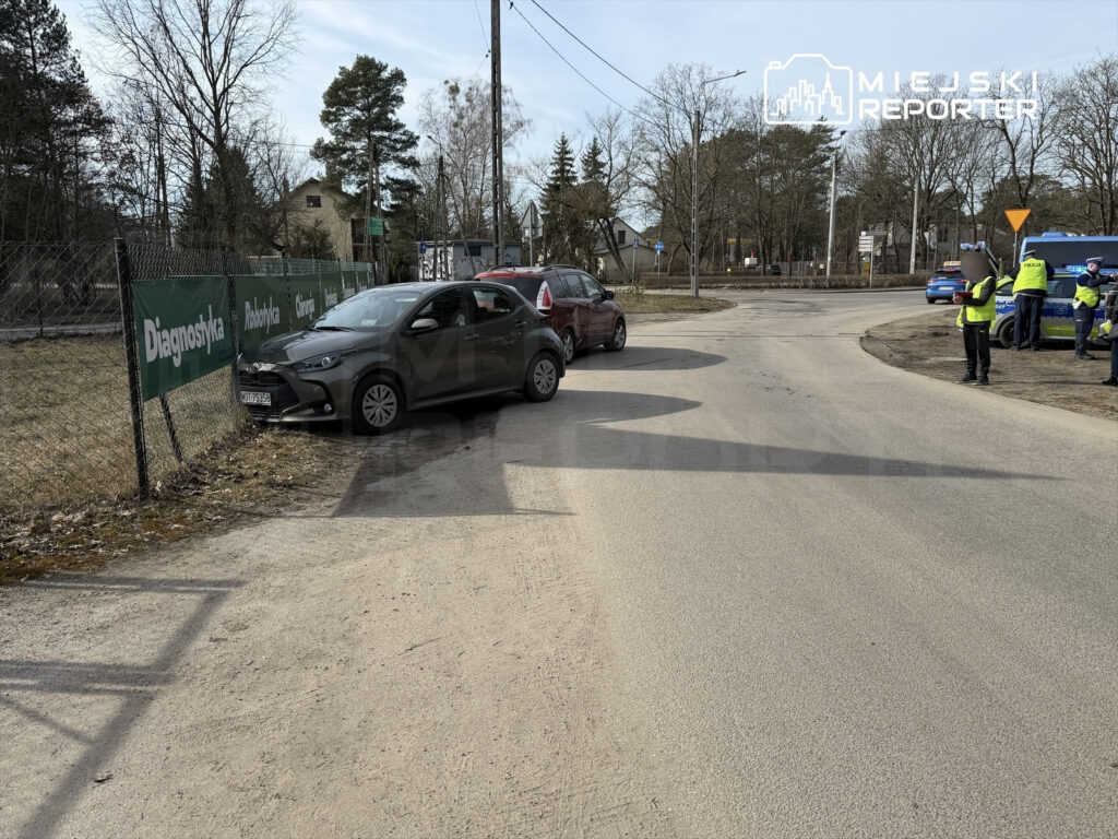 Dwa samochody zaparkowane na poboczu drogi, obok ogrodzenia z reklamami, w tle policjanci rozmawiający z kierowcami.