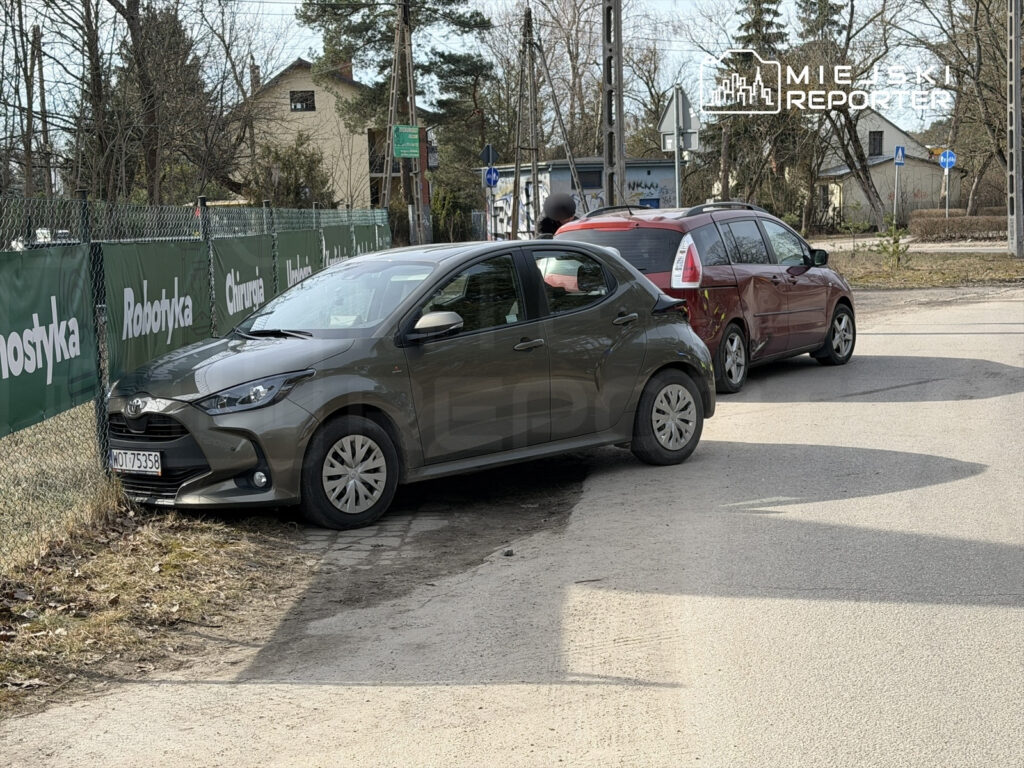 Szary samochód zaparkowany blisko ogrodzenia, obok czerwonego auta, w otoczeniu drzew i reklam.