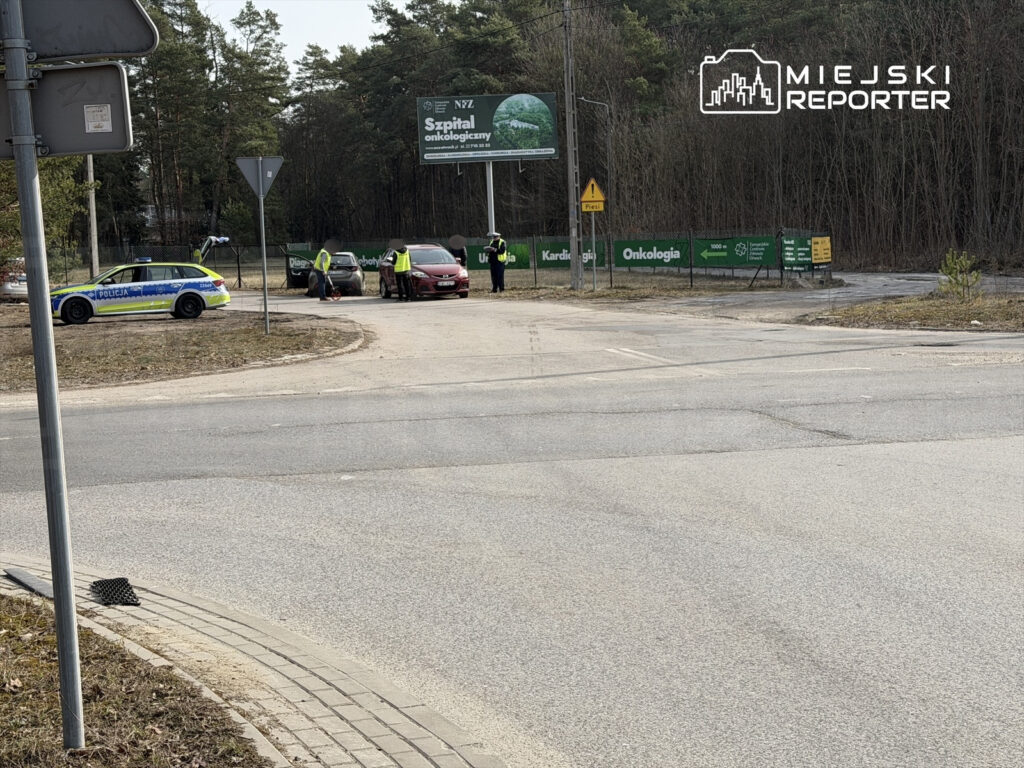 Policjanci kontrolujący pojazdy na skrzyżowaniu w pobliżu ogrodzenia szpitala onkologicznego.