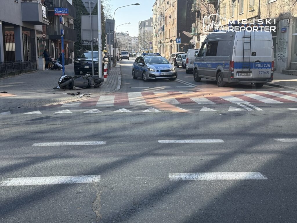 Na ulicy leży przewrócony skuter, obok zatrzymane pojazdy policyjne i osobowe, w tle widać budynki mieszkalne.
