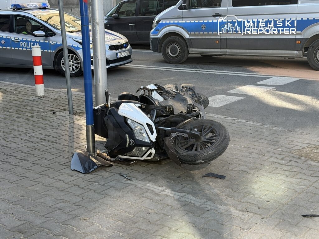 Motocykl leży na boku na chodniku obok słupa, a w tle widoczny radiowóz policyjny.