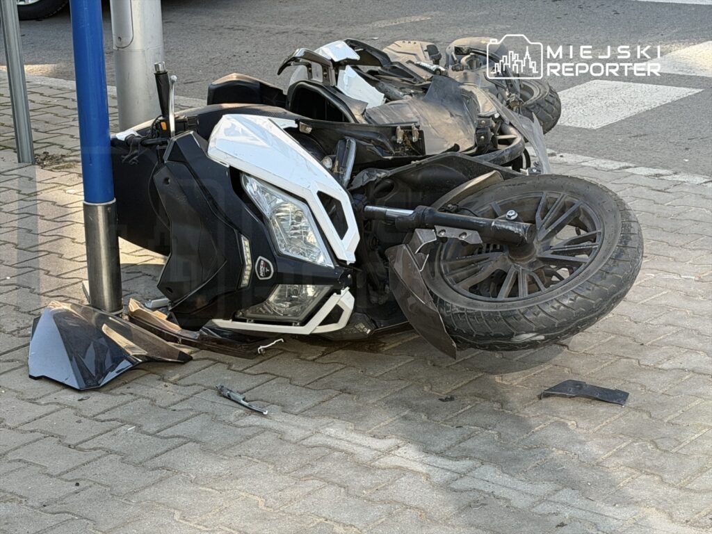 Złamany skuter leży na boku obok słupa, z uszkodzoną osłoną i odrzuconymi elementami na chodniku.