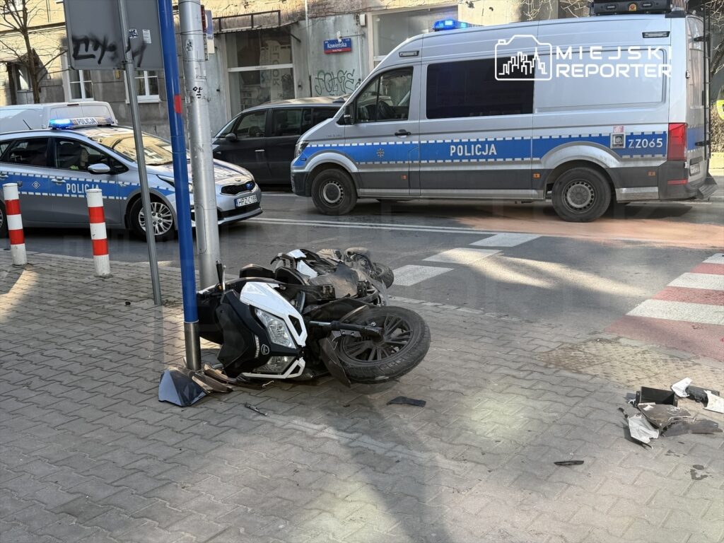 Motocykl leży przewrócony na chodniku obok policyjnych radiowozów w miejskim otoczeniu.