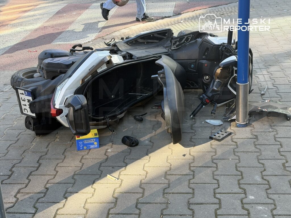 Przewrócony skuter leży na chodniku obok słupa, z widocznymi uszkodzeniami i częściami rozrzuconymi wokół.