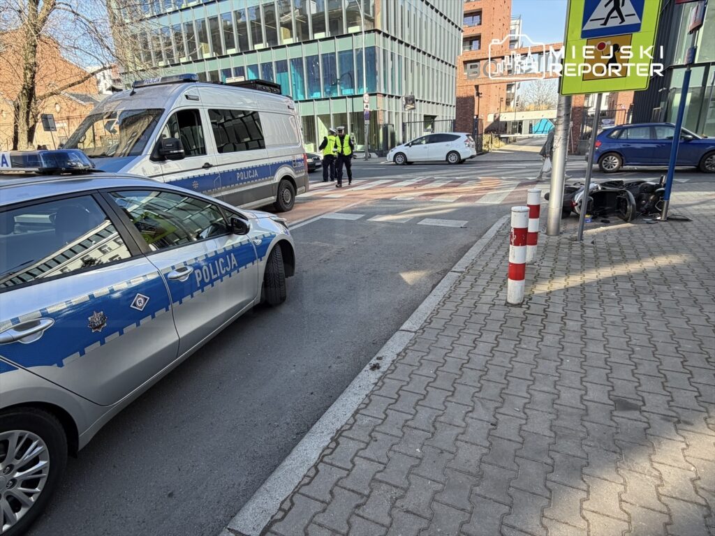 Policyjny radiowóz i furgonetka zatrzymane na skrzyżowaniu, obok policjanci kontrolujący ruch. W tle nowoczesne budynki.