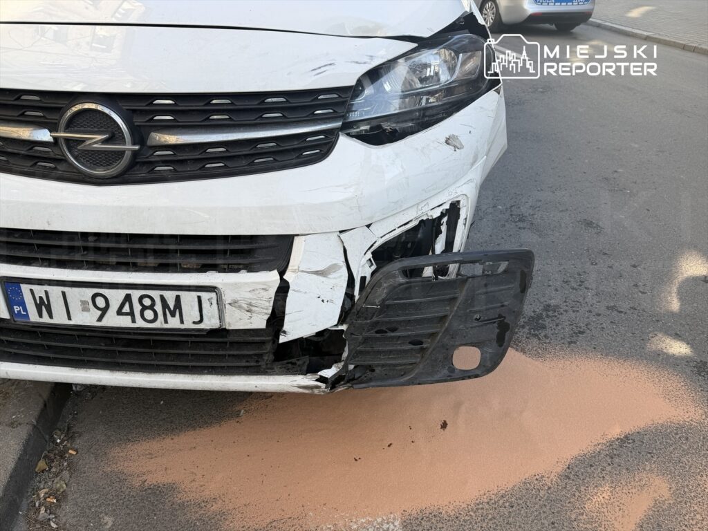 Uszkodzony biały samochód osobowy z widocznym uszkodzeniem przedniego zderzaka stoi na ulicy, obok leży różowy proszek.