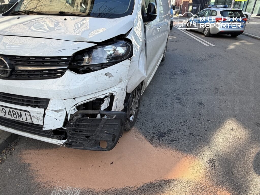 Rozbity biały van zaparkowany na ulicy, z uszkodzoną przednią częścią i plamą różowego płynu na jezdni.