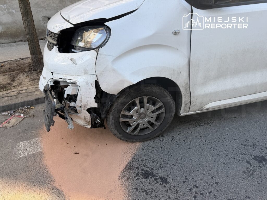 Uszkodzony biały samochód dostawczy z otwartą maską, stojący na ulicy z plamą piasku pod przednim zderzakiem.