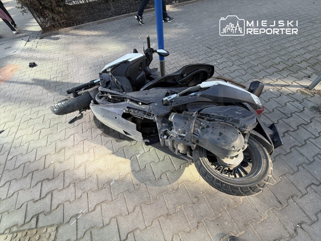 Zniszczony skuter leżący na chodniku, z widocznymi uszkodzeniami i przewróconym kołem, obok przechodzący piesi.