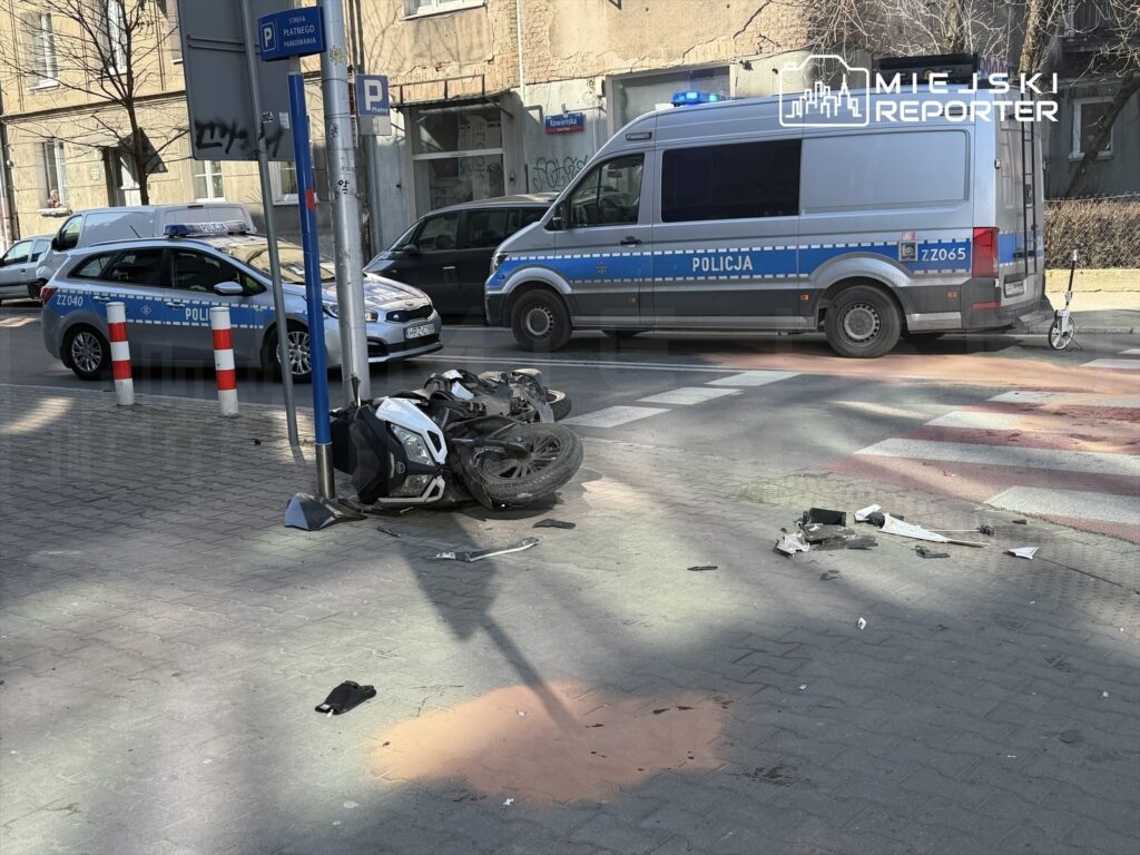 Motocykl leży przewrócony na chodniku, obok stoją policyjne pojazdy oraz rozrzucone fragmenty.