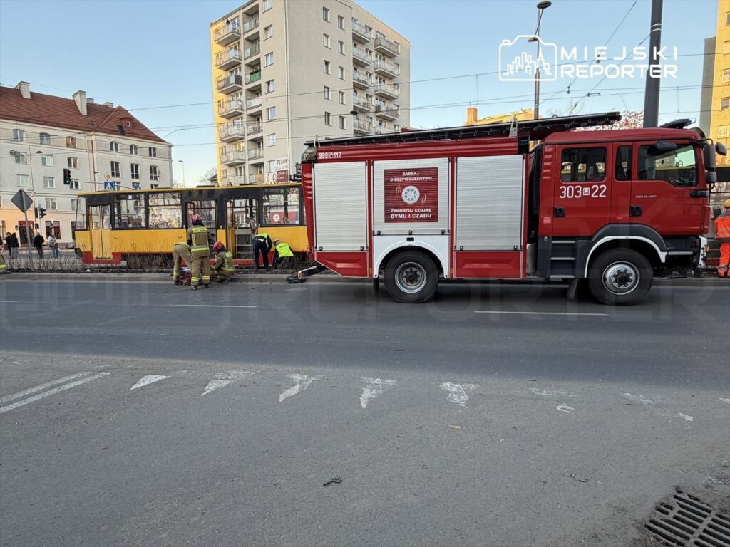 Strażacy w mundurach pracują przy uszkodzonym tramwaju, obok stoi wóz strażacki na ruchliwej ulicy.