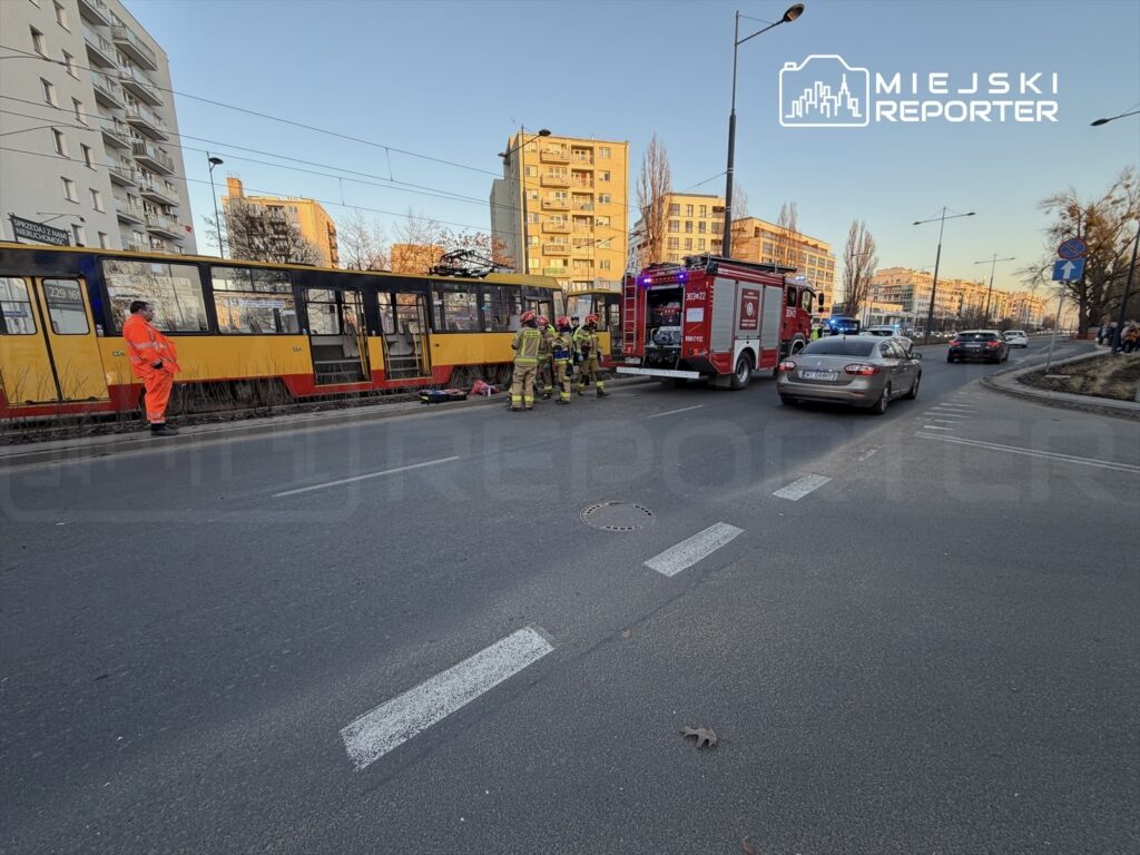 Zespół ratunkowy w akcji obok tramwaju, przy drodze w miejskim otoczeniu, z widocznym pojazdem straży pożarnej.