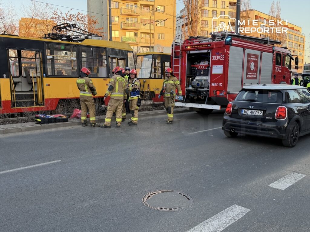 Grupa strażaków w hełmach omawia sytuację obok uszkodzonego tramwaju, obok stoi wóz strażacki.