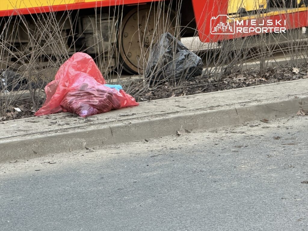 Czerwony worek na śmieci leży na chodniku obok czarnego worka, w tle widoczne koła pojazdu.