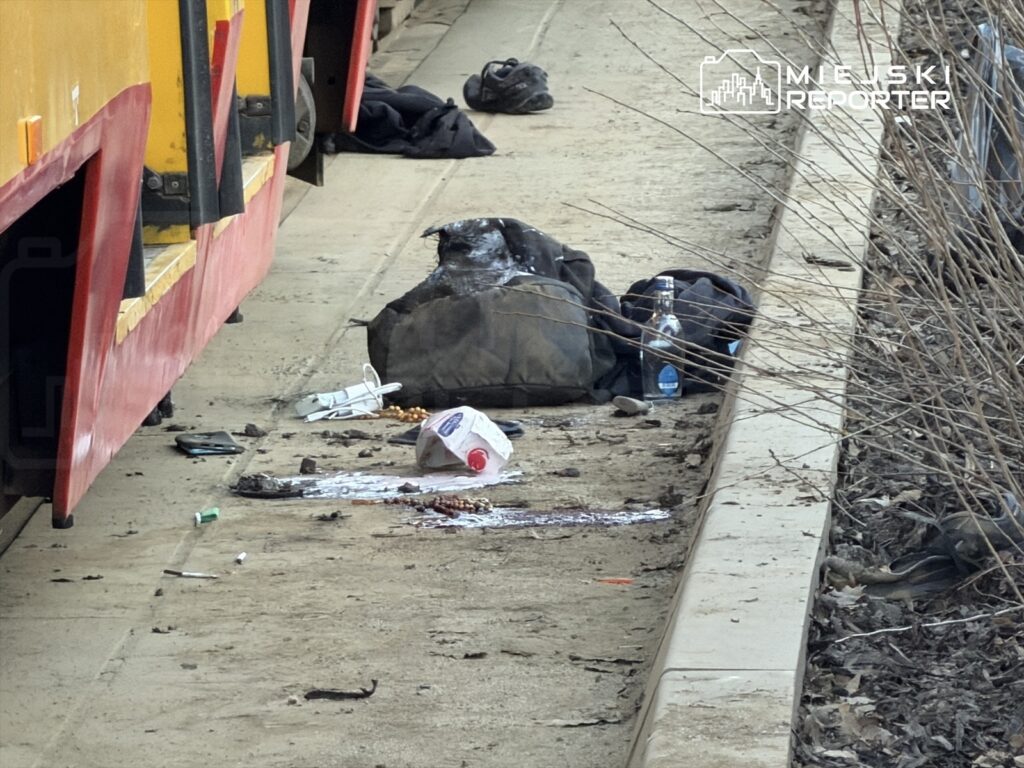 Na peronie leżą porzucone plecaki, butelka wody oraz odpady, w tle czerwony pojazd komunikacji miejskiej.