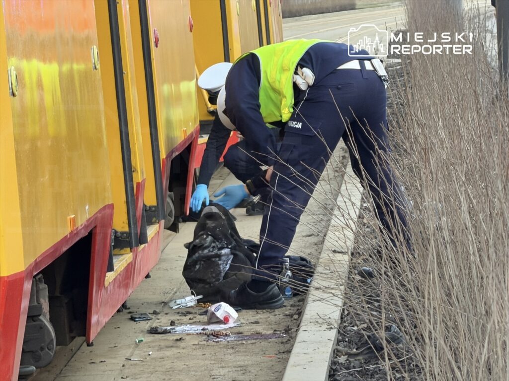 Funkcjonariusze policji przeszukują torowisko obok tramwaju, zbierając porzucone przedmioty.