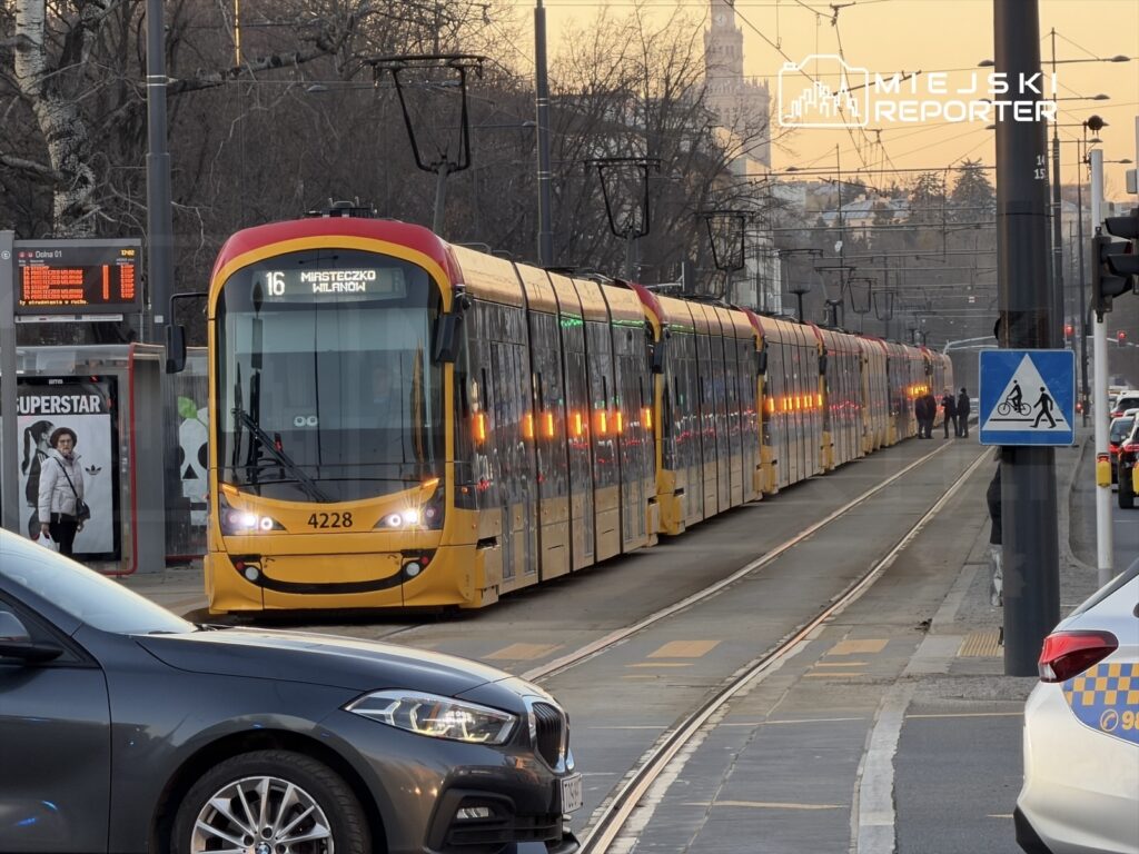 Tramwaj linii 16 zatrzymany na przystanku, obok czekają pasażerowie, a w tle widoczne są samochody i budynki.
