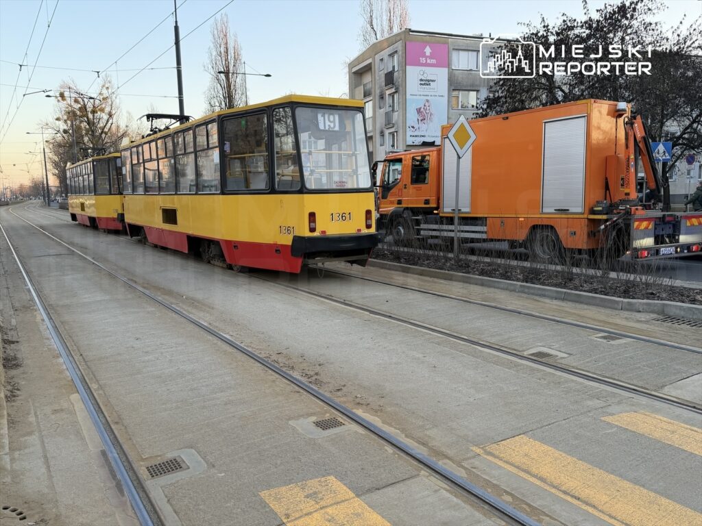 Tramwaj w żółto-czerwonym malowaniu zatrzymany na torowisku obok pomarańczowego pojazdu serwisowego.