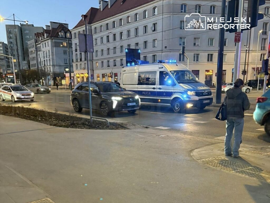 Radiowóz policyjny z włączonymi sygnałami świetlnymi przejeżdża obok ruchliwej ulicy, obok pieszy z torbą.