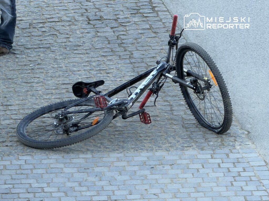 Czarny rower górski leżący na kostce brukowej, z czerwonymi pedałami i widocznymi oponami.
