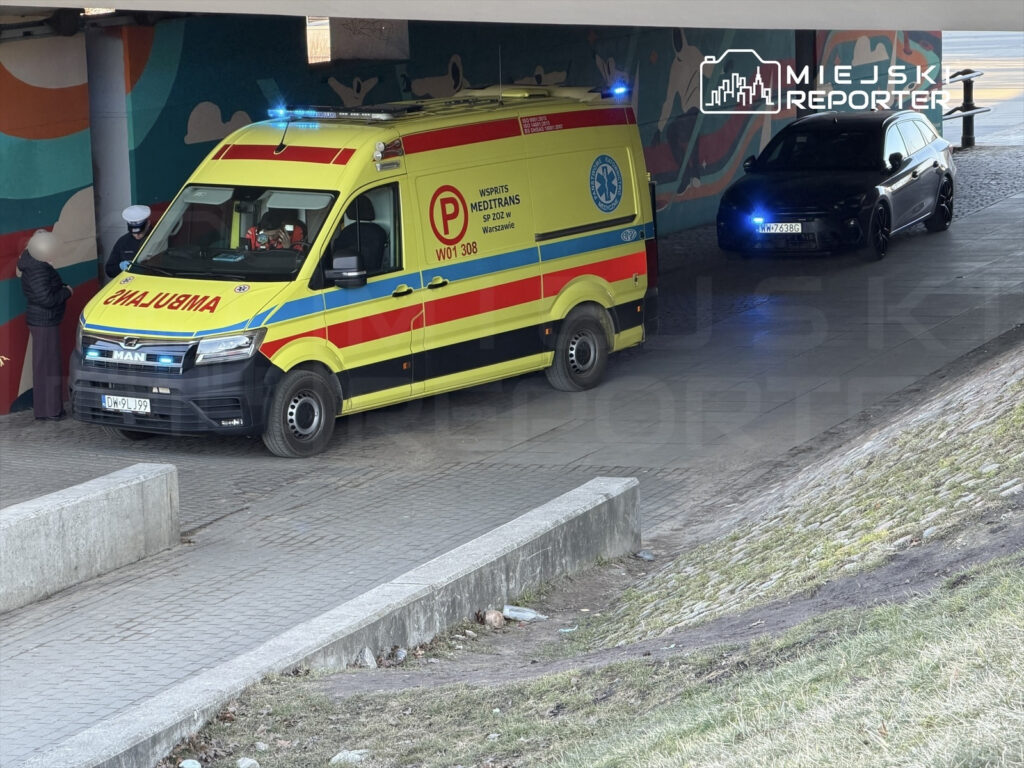 Karetka pogotowia z włączonymi sygnałami świetlnymi stoi pod zadaszeniem, obok niej policjant rozmawia z osobą w ciemnym ubraniu.