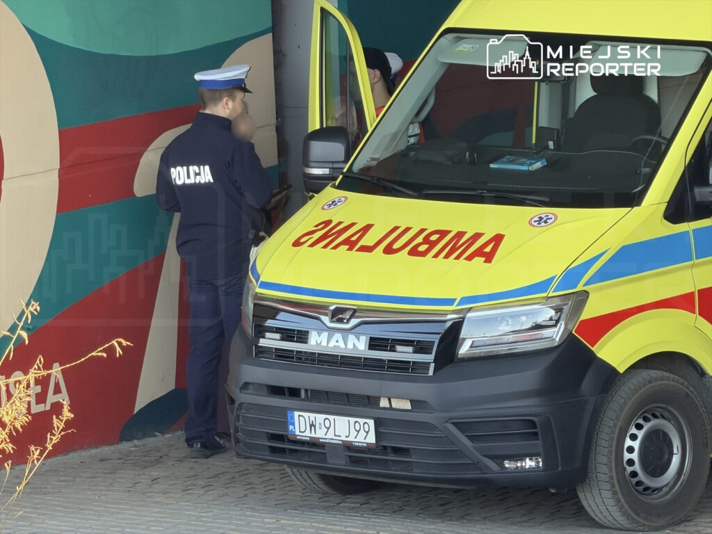 Policjant rozmawia z ratownikiem medycznym przy ambulansie w kolorze żółto-czerwonym, w tle mural.