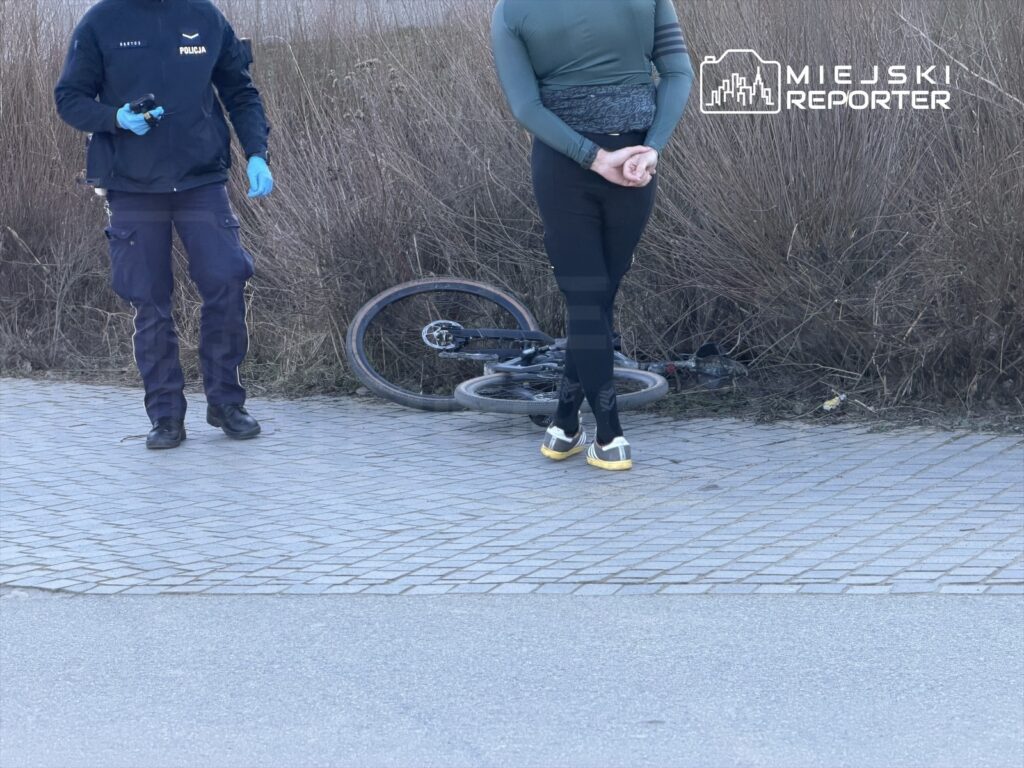 Policjant z bronią w ręku stoi obok leżącego roweru na chodniku, a obok niego mężczyzna w obcisłym ubraniu.