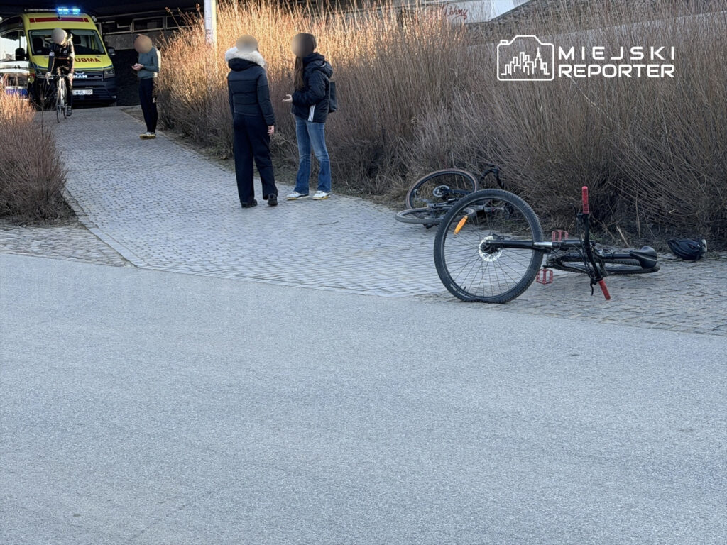 Grupa osób rozmawia obok przewróconych rowerów na ścieżce, w tle widoczny ambulans.