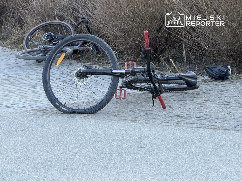 Na ścieżce rowerowej leży przewrócony rower górski z czerwoną kierownicą i kask. W tle widać drugi rower.