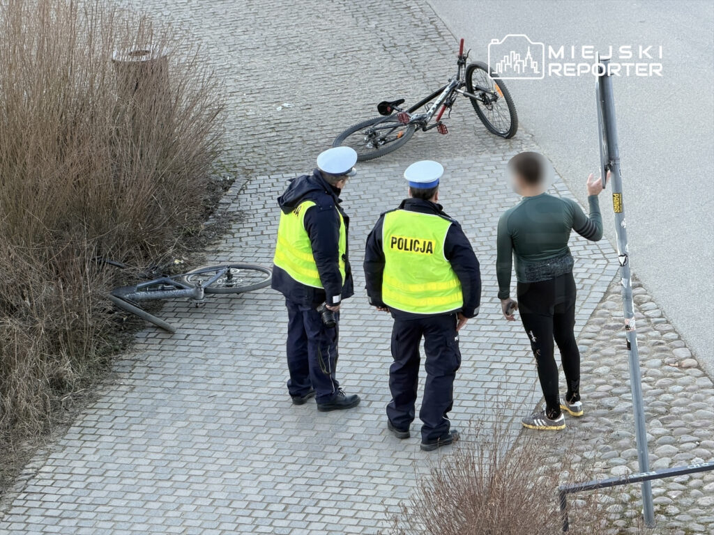 Mężczyzna w obcisłej odzieży rozmawia z dwoma policjantami w kamizelkach odblaskowych obok przewróconego roweru.