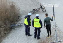 Mężczyzna w obcisłej odzieży rozmawia z dwoma policjantami w kamizelkach odblaskowych obok przewróconego roweru.