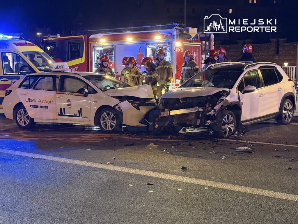 Dwa uszkodzone samochody osobowe, w tym taksówka z napisem "Uber Airport", na miejscu wypadku, otoczone przez strażaków i służby ratunkowe.