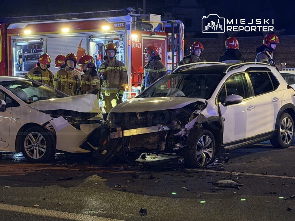 Grupa strażaków w hełmach omawia sytuację przy zderzeniu dwóch uszkodzonych samochodów na ulicy nocą.